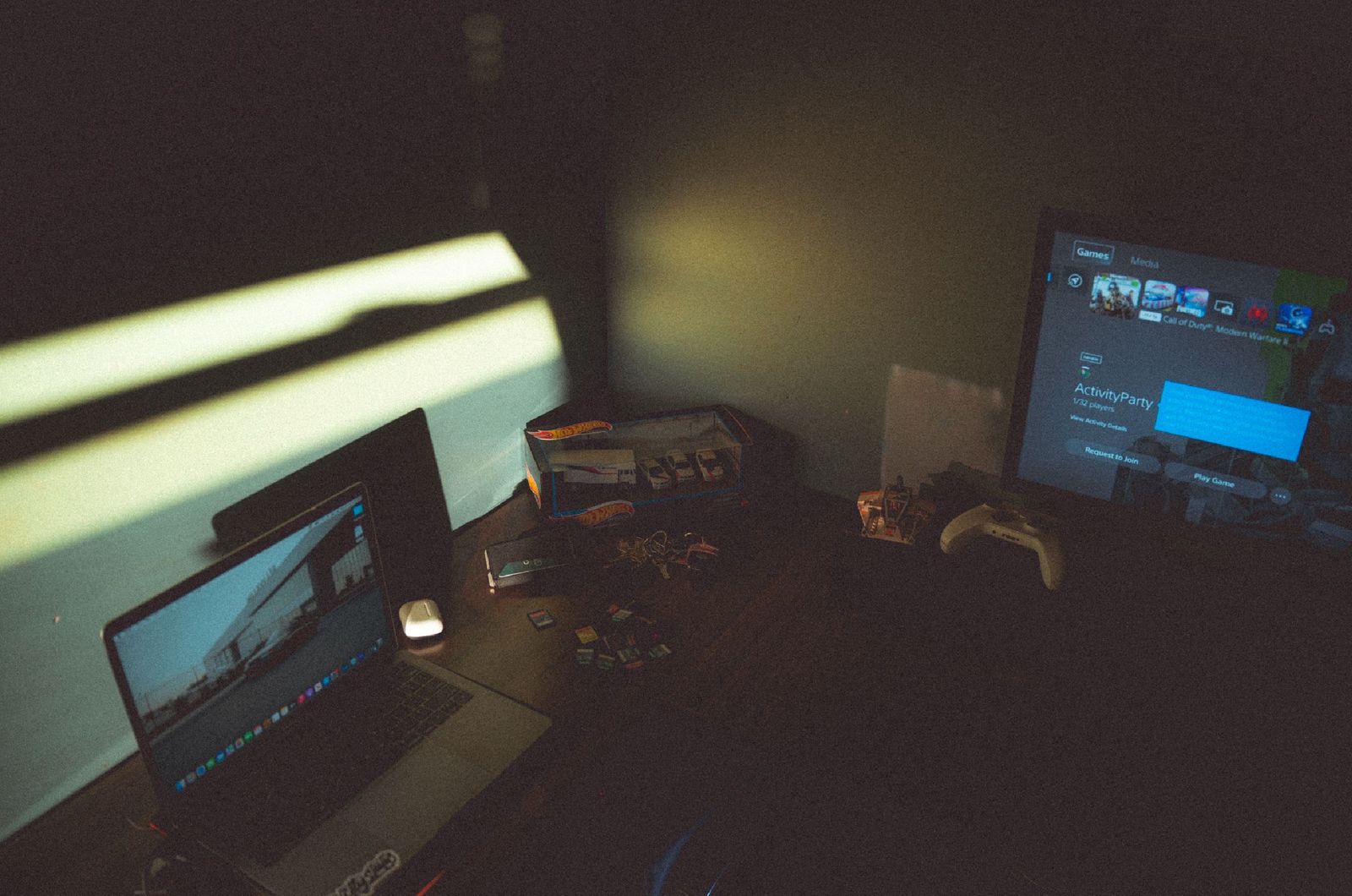 A young man playing a video game on a computer with a tired expression, his work laptop open beside him.